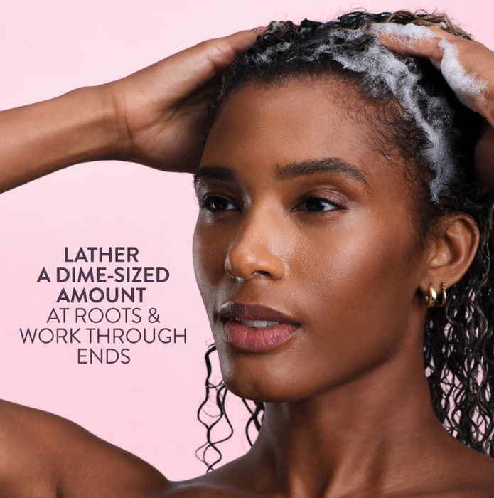 A close-up of a woman with wet, curly hair covered in lathered shampoo foam, set against a solid pink background with promotional text on the left.